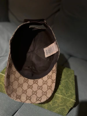 Gucci materialmix keps med GG-mönster - Säljer en snygg beige och brun keps från Gucci med klassiskt GG-monogram över hela kepsen. Kepsen har justerbart band bak i brunt läder och en ikonisk röd-grön rand på sidan. Materialmix i bomull och polyester, perfekt för dig som gillar exklusiva accessoarer.