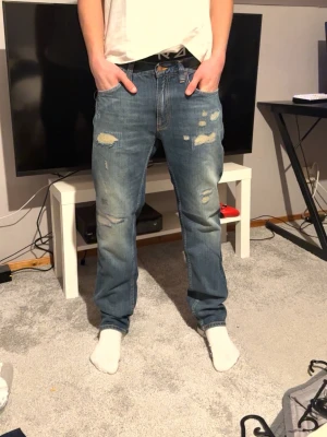 Nudie Jeans blå slitna jeans - Säljer ett par blå jeans från Nudie Jeans med slitna detaljer och lagningar på benen. orange sömmar och är gjorda i ekologisk bomull. Passformen är rak och de har en normal midja. Perfekta för en avslappnad och cool stil. Jeansen är helt nya lappar är kvar. W29/L30 priset är ej hugget i sten 