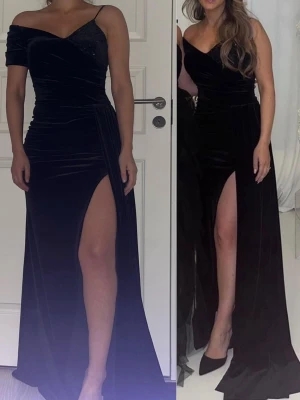 Svart sammetsklänning med slits - Elegant svart långklänning i mjuk sammet med en djup slits på ena sidan. Klänningen har omlott-v-ringning, en axelbandslös sida och en sm logg axelrem på andra sidan. S wholett detaljer upptill ger extra lyxig känsla.