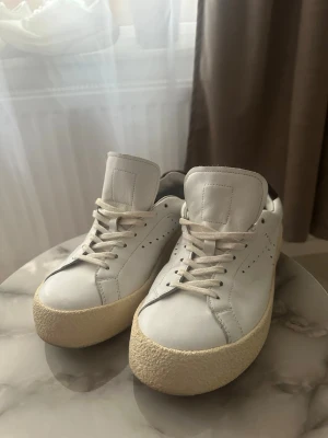Vita Kenzo sneakers med beige sula - Säljer ett par vita Kenzo sneakers i skinn med beige, grov platt sula och diskreta håldetaljer på sidorna. Skorna har snörning och mörklila detalj på hälen. Klassisk och clean look som passar till allt.