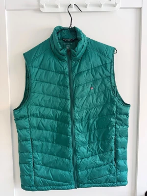 Grön dunväst från GANT - Snygg grön dunväst från GANT med quiltad design och hög krage. Västen har dragkedja framtill, två sidofickor och en liten GANT-logga på bröstet. Perfekt att slänga över en hoodie eller tröja för extra värme.