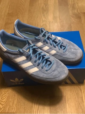 Adidas Handball Spezial ljusblå mocka - Säljer ett par Adidas Handball Spezial sneakers i ljusblå mocka med vita detaljer och klassiska tre ränder på sidan. Skorna har snörning, brun yttersula och retroinspirerad design. Perfekta för dig som gillar streetwear och sportig stil. Ställ fråga vid minsta fundering 🤔. Pris kan såklart diskuteras vid snabb affär