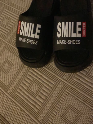 Svarta slides med text SMILE - Svarta slides med bred rem där det står 'SMILE SURPER MAKE-SHOES' i vit och röd text. Skorna har öppen tå och platt sula, perfekta för sommaren. Tillverkade i syntetmaterial och har en enkel, clean look.