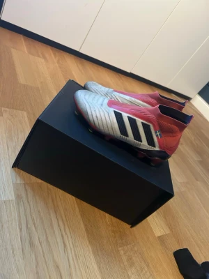 Adidas Predator 18+ SG fotbollsskor - Säljer ett par Adidas Predator 18+ SG fotbollsskor i vitt, svart och rött. Skorna har en strumpliknande ovandel i syntetmaterial och klassiska svarta ränder på sidan. Perfekta för dig som vill sticka ut på planen och få grym kontroll på bollen. Skorna går att få renare har bara inte haft orken till att göra det, skruv dobb kommer till men lite nu är det ett par skruvdobbar som är lite mindre vassa och inte i metall.Kan sänka priset! Orginal pris:2599 
