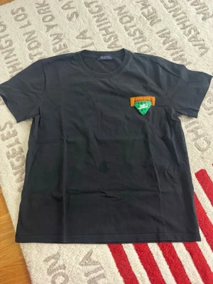 Svart Louis Vuitton t-shirt med stor LV-logga - Säljer en svart t-shirt från Louis Vuitton med stor vit och grön LV-logga på ryggen och en grön patch med logga på bröstet. Klassisk rund hals och korta ärmar. T-shirten är i mjuk bomull och har bruna detaljer med Louis Vuitton-tryck.