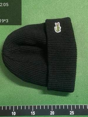 Svart ribbad mössa från Lacoste - Säljer en svart ribbstickad mössa från Lacoste med den klassiska gröna krokodilloggan framtill. Mössan är enkel och stilren med vikt kant och passar perfekt till kyliga dagar. Den också helt ny