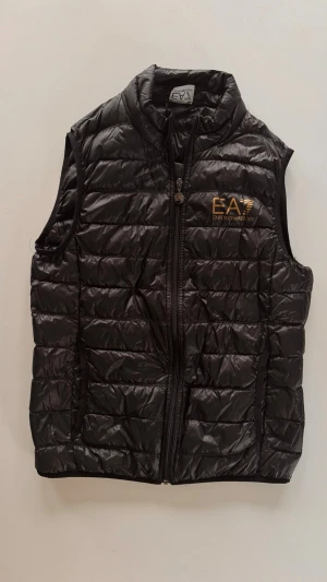 Svart dunväst från Emporio Armani - Snygg svart dunväst från Emporio Armani EA7 med guldfärgad logga på bröstet. Västen har quiltad design, hög krage och dragkedja framtill. Perfekt att slänga över en hoodie eller tröja för en sportig och clean look. Storlek S.           Använd ett sommarlov 