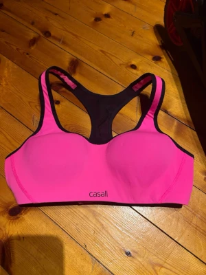 Rosa sport-bh från Casall Sculpture - Sport-bh från Casall Sculpture i stark rosa med svarta detaljer. Modellen har vadderade kupor, breda axelband och justerbar knäppning bak. Materialet är mjukt och stretchigt syntet, perfekt för träning och hög komfort.