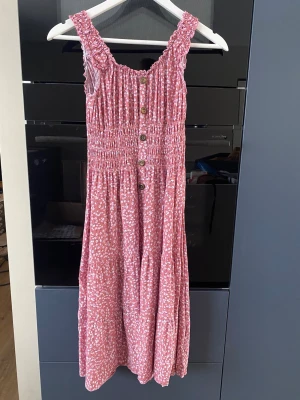 Rosa blommig långklänning med knappar - Supersöt långklänning i rosa med små vita blommor och dekorativa knappar framtill. Klänningen har breda axelband med volangkant och smockad överdel som ger en bekväm passform. Perfekt för varma dagar och somriga vibes!