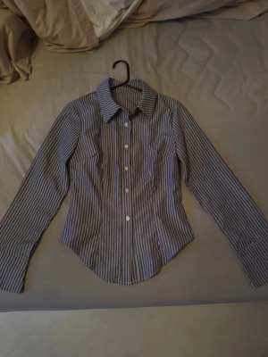 Randig blåvit skjorta med knappar - Snygg blåvit skjorta med smala vertikala ränder och klassisk krage. Skjortan har långa ärmar, vita knappar framtill och markerad midja för en schysst passform. Perfekt till jeans eller kjol för en clean look.