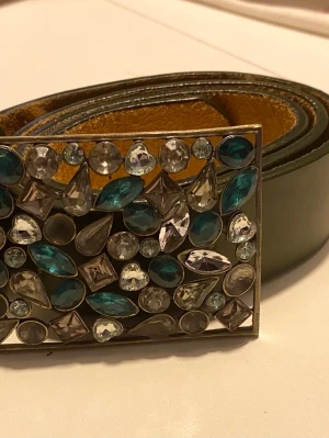 Grönt bälte med stenar  - Grönt bälte med ett rektangulärt spänne dekorerat med färgade och klara strasstenar i olika former. Skinnimitation 105 X 3,5cm Spänne: 9x6 cm Obs, det fattas pärlor!