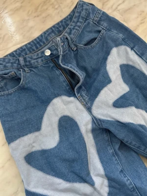 Blå jeans med vita hjärtan - Unika blå jeans med stora vita hjärtan målade över hela byxbenen. De har en rak passform och klassisk femficksdesign. Perfekta för dig som vill sticka ut och addera lite kärlek till din outfit.