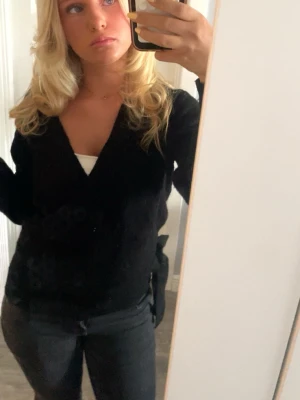 Svart omlottkofta från Liz Devy - Säljer en svart omlottkofta från Liz Devy i storlek S. Koftan har v-ringning och långa ärmar, perfekt att slänga på sig över ett linne. Enkel och stilren design som funkar till många outfits.