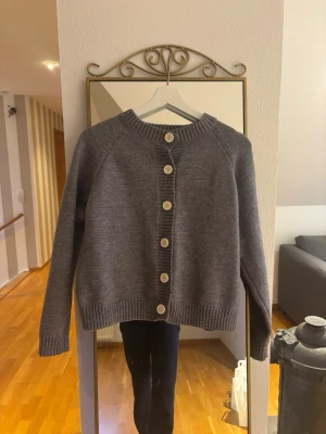 Grå kofta från Design by Amor - Mysig grå kofta från Design by Amor med grov ribbstickad kant och stora vita knappar framtill. Koftan har lång ärm och en rak, något boxig passform som ger en chill vibe. Perfekt att slänga över en t-shirt eller linne när det blir kyligt.