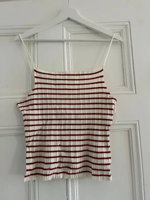 Randig ribbad linnetopp från H&M - Säljer en vit ribbad linnetopp från H&M med tunna axelband och röda horisontella ränder. Toppen är stretchig och har en avslappnad passform. Axlebanden är tyvärr väldigt långa och åker ner på mig. Tveka inte på att höra av dig om du har frågor❤️