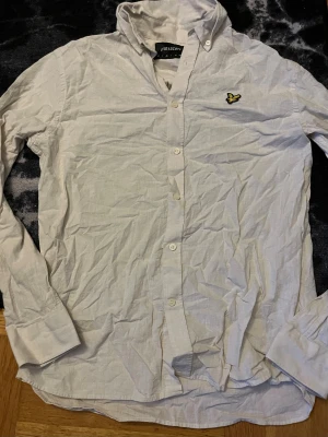 Vit skjorta från Lyle & Scott, M - Klassisk vit skjorta från Lyle & Scott i storlek M. Skjortan har lång ärm, knappar framtill och den ikoniska broderade gula örnen på bröstet. Tillverkad i ett lätt och svalt linnematerial, perfekt för en clean och stilren look.