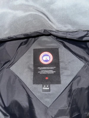 Svart pufferjacka från Canada Goose - Säljer en svart pufferjacka från Canada Goose med huva och klassisk logga på ärmen. Jackan har dragkedja, stora fickor framtill och ribbade muddar vid ärmsluten. Perfekt för kalla dagar och riktigt snygg till streetwear-stilen.