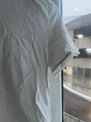 Vit topp med spetskant från Zara - Säljer en enkel vit topp från Zara i storlek S. Toppen har rund halsringning, korta ärmar och fina spetsdetaljer längst ut på ärmsluten. Tillverkad i mjuk bomull som känns skön mot huden. Perfekt att styla med jeans eller kjol.