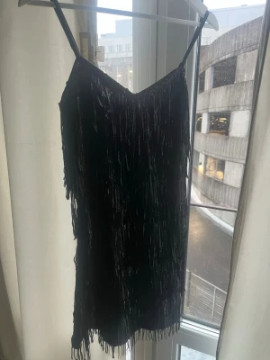 Svart glittrig fransklänning - Säljer en svart kort klänning med tunna axelband och massor av glittriga fransar som ger en riktigt festlig känsla. Klänningen har en rak passform och är täckt av paljetter som reflekterar ljuset snyggt. Perfekt för dig som vill sticka ut på nästa fest!