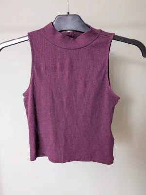 Mörklila ribbad croppad polotopp - Säljer en mörklila croppad polotopp i ribbad bomullsmix. Toppen är ärmlös och har en hög krage som ger en clean och trendig look. Perfekt att styla med höga jeans eller kjol för en chill vibe.