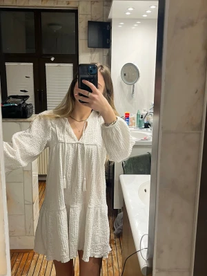 Vit klänning med volang - Superfin vit klänning med volangkant och långa ärmar. Klänningen har struktur i tyget, knytband med tofsar vid halsen och en luftig passform. Perfekt för varma dagar och ger en avslappnad men söt look.