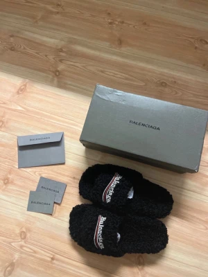Balenci fur slides - Säljer ett par svarta balenci fur slides i mjukt teddy-material. De har öppen tå, platt sula och broderad logga med vit och röd detalj på ovandelen. Perfekta för chill dagar hemma eller när du vill ha något extra cozy på fötterna.