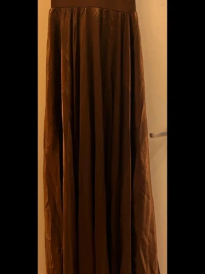 Brun långklänning med slits - Säljer en brun långklänning i glansigt material med smala axelband. Det är resår i ryggen så det är stretchigt runt bysten. Klänningen har en snygg slits framtill. Perfekt för dig som vill ha en elegant look med en twist.