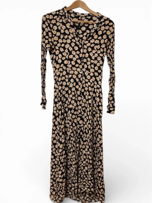 Blommig långklänning från Ganni - Supersnygg långklänning från Ganni med svart botten och beige blommönster. Klänningen har långa ärmar, v-ringning och knappar framtill. Tillverkad i mjuk viskos som ger ett fint fall. Perfekt för dig som vill ha en cool och trendig look.