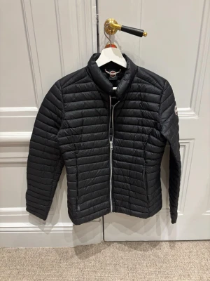 Svart pufferjacka från Colmar - Säljer en svart pufferjacka från Colmat med horisontella quiltade sömmar och dragkedja framtill. Jackan har hög krage och en clean, sportig look. Perfekt för dig som vill ha en lätt och smidig jacka till vardags.