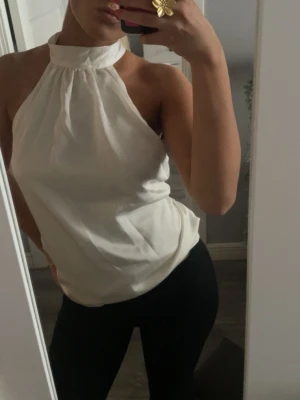 Vit halterneck topp från Amisu - Säljer en snygg vit halterneck topp från Amisu i blankt, mjukt material. Toppen har hög krage och är ärmlös med öppen rygg, vilket ger en clean och modern look. Perfekt till sommarens outfits och lätt att styla med både jeans och kjol.