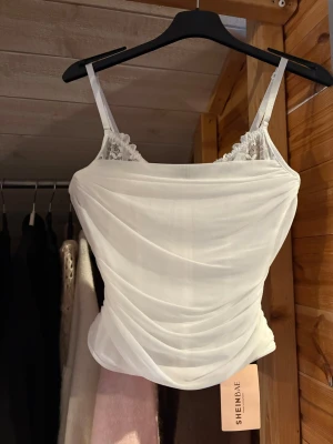 Vit korsettopp med spets från SHEIN - Säljer en vit korsettopp från SHEIN med tunna axelband och fina spetsdetaljer vid bysten. Toppen har draperad mesh över hela framsidan och baksidan, samt en dold dragkedja i ryggen. Perfekt för dig som gillar romantiska och trendiga plagg.