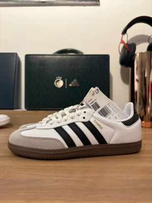 Adidas Samba OG vit/svart 37 1/3 - Adidas Samba OG sneakers i vitt skinn med svarta detaljer och klassiska tre ränder på sidan. Grå mockadetaljer vid tån och brun gummisula. Guldtexten 'SAMBA' på sidan och snörning framtill. Perfekt för dig som gillar retrostil och streetwear.( Äkta morsan jobbar där) Aldrig andvänd