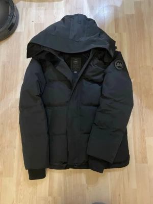Canada Goose Black Label Mcmillian - Säljer en svart pufferjacka från Canada Goose med stor huva och patch på ärmen. Jackan har långa ärmar, dragkedja och knappar framtill. Perfekt för kalla dagar och riktigt snygg till vinterlooken.
