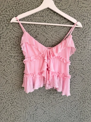 Rosa volangig linnetopp med snörning - Supersöt rosa linnetopp med tunna axelband och volangdetaljer. Toppen har smala vertikala ränder, snörning framtill och flera lager volanger som ger en romantisk vibe. Materialet är lätt och känns luftigt, perfekt för varma dagar.