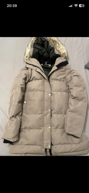 Beige pufferjacka med fuskpäls - Säljer en beige pufferjacka från Everest i modellen W pelona down coat.  med avtagbar huva och fuskpälsdetalj. Jackan har långa ärmar, dragkedja och knappar framtill samt flera praktiska fickor. Perfekt för kalla dagar och riktigt mysig att bära. Pris kan diskuteras!