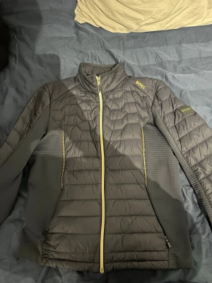 Svart dunjacka från BOSS med gul dragkedja - Snygg svart dunjacka från BOSS med gul dragkedja och detaljer. Jackan har quiltad framsida och baksida, hög krage och två sidofickor. Lättviktig och vattenavvisande, perfekt för kyliga dagar. BOSS-logga på axeln och ärmen för extra stilpoäng.