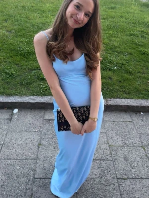 Ljusblå långklänning med öppen rygg - Supersnygg ljusblå långklänning med smala axelband och öppen rygg med dekorativa band. Klänningen har en figurnära passform och är tillverkad i ett glansigt, slätt material som ger en lyxig känsla. Perfekt för sommarens speciella tillfällen!