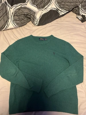 Grön stickad tröja från Polo Ralph Lauren - Mysig grön stickad tröja från Polo Ralph Lauren med klassisk rund halsringning och broderad logga på bröstet. Tröjan har långa ärmar och ribbade muddar vid ärmslut och nederkant. Perfekt för lager-på-lager under kyliga dagar.tröjan har ett väldigt litet hål precis vid den högra armhålan men det krävs nog inte mer en ett par stygn för att laga de:) pris ej hugget i sten