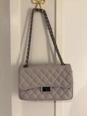 Ljusgrå quiltad axelväska - Snygg ljusgrå axelväska med quiltat mönster och kedjerem. Väskan har en klassisk rektangulär form och stängs med ett metallspänne framtill. 💕