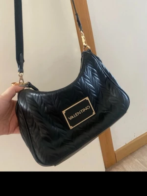 Svart quiltad axelväska Valentino - Säljer en svart quiltad axelväska från Valentino med snyggt mönster och guldfärgade detaljer. Väskan har ett stort metallogo framtill, justerbar axelrem med logotyp och dragkedja upptill. Perfekt för dig som vill ha en trendig och praktisk väska.