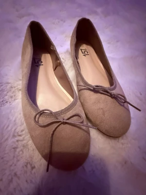 Beige ballerinaskor med rosett från 157 - Säljer ett par stilrena beige ballerinaskor från 157 i storlek 36. Skorna har en mjuk mockaliknande yta, rund tå och en söt rosettdetalj framtill. Sulan har hjärtformade mönster för extra grepp. Perfekta för dig som gillar enkel och klassisk stil.