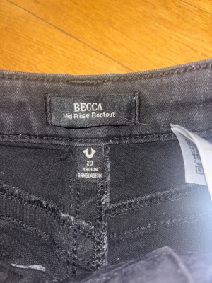 Svarta bootcut jeans True Religion - Svarta jeans från True Religion, modell BECCA Mid Rise Bootcut. Klassisk femficksdesign med snygga detaljer och knappar på bakfickorna. Jeansen har medelhög midja och bootcut-ben, tillverkade i stretchigt jeansmaterial för en bekväm passform.