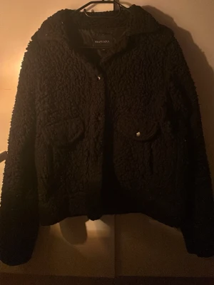 Svart teddyjacka från Brave Soul - Mysig svart teddyjacka från Brave Soul med två fickor framtill och knappar. Jackan har en fluffig struktur och är perfekt för kyliga dagar. Enkel och stilren design som passar till många olika outfits.