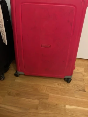 Röd resväska från Samsonite - Stor och rymlig röd resväska från Samsonite med fyra hjul och praktiskt handtag. Väskan har en stilren design i hårdplast och är perfekt för längre resor. Enkel att rulla och packa med smart lås upptill.
