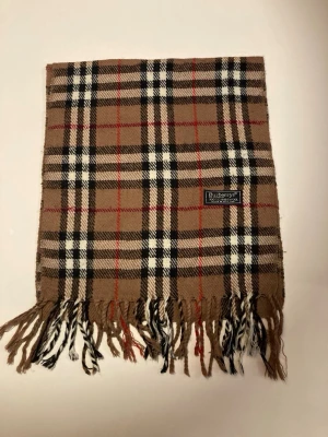 Rutig halsduk från Burberrys i ull - Vi säljer denna otroligt snygga burberry halsduk med färgerna beige, röd, svart och vit. Utan defekter