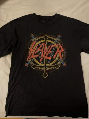 Svart Slayer t-shirt - Svart Slayer t-shirt med stort tryck fram i rött, gult och blått. Klassisk rund hals och loose fit. Tillverkad i mjuk bomull och har korta ärmar. Tröjan är tunn och skön! Sitter ungefär som en S!