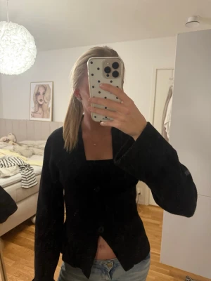 Svart kofta med knappar - Säljer en svart kofta med två stora knappar framtill och breda ärmar. Koftan har en djup v-ringning och är lätt croppad. Perfekt att slänga över ett linne eller t-shirt för en chill look.