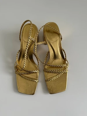 Guldiga sandaler med flätade remmar från Zara - Säljer ett par guldiga sandaler från Zara med fyrkantig tå och flätade remmar. Skorna har en låg klack och öppen tå, vilket ger en trendig och elegant look. Tillverkade i syntetmaterial med metallic-finish och stilrena detaljer.
