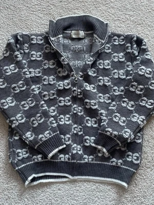 Gucci cardigan grå - Säljer en grå kofta från Gucci med hel dragkedja och ribbad krage. Tröjan har ett upprepat GG-mönster i vitt och grått, samt ribbade muddar och nederkant. Perfekt för dig som gillar exklusiva och snygga plagg med tydlig logga. Koftan är i storlek M men passar mer som S. Tveka inte att höra av er för mer frågor eller bilder🤗