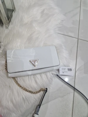 Vit axelväska Guess med kedja - Supersnygg vit axelväska från Guess med guldfärgad kedja och klassisk trekantslogga framtill. Väskan har struktur i materialet, flera kortfack och dragkedja inuti. Perfekt storlek för det viktigaste och stilren look.